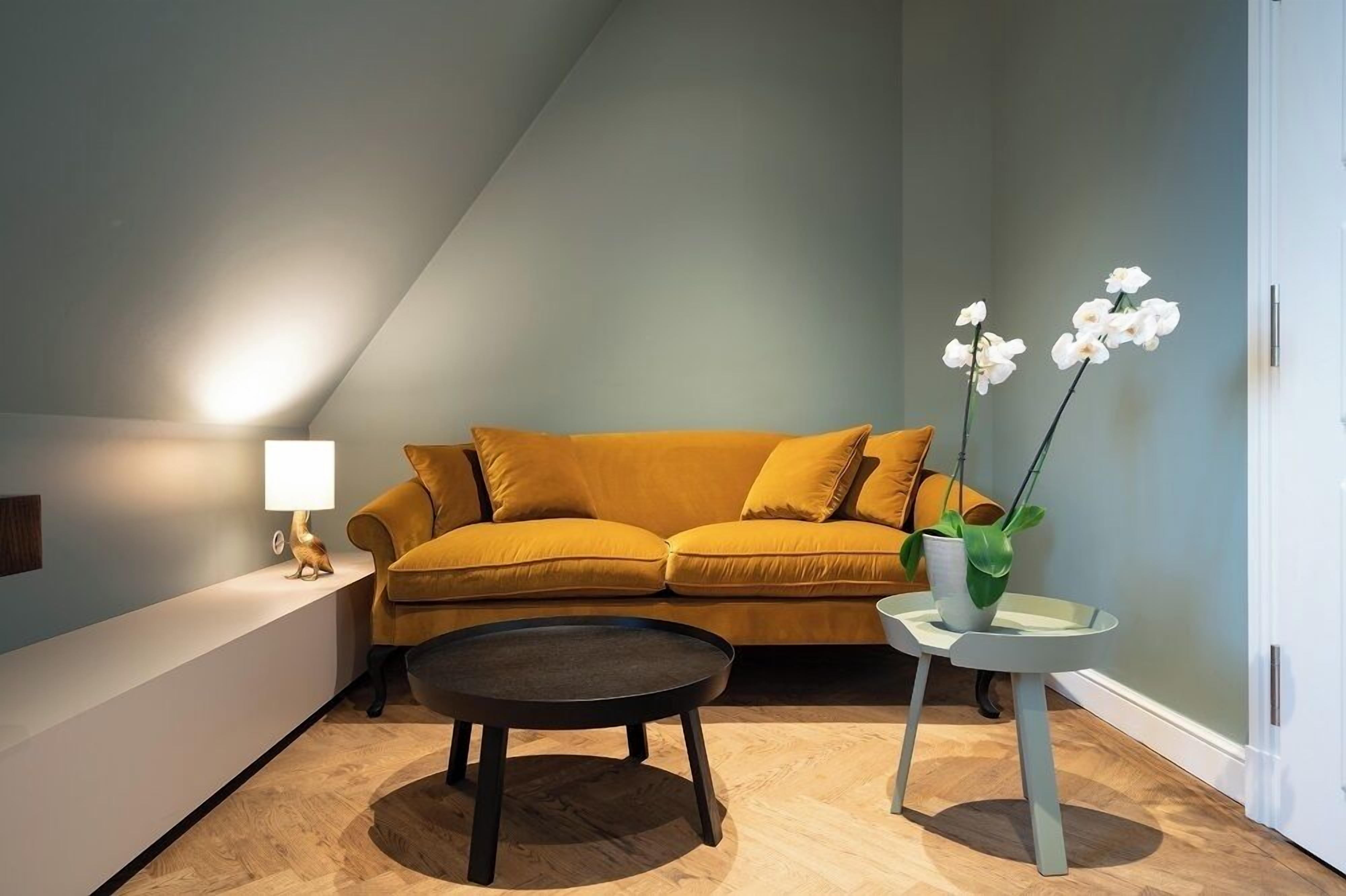 Een moderne woonkamer met een mosterdgele bank, een klein rond tafeltje en een orchidee in pot.