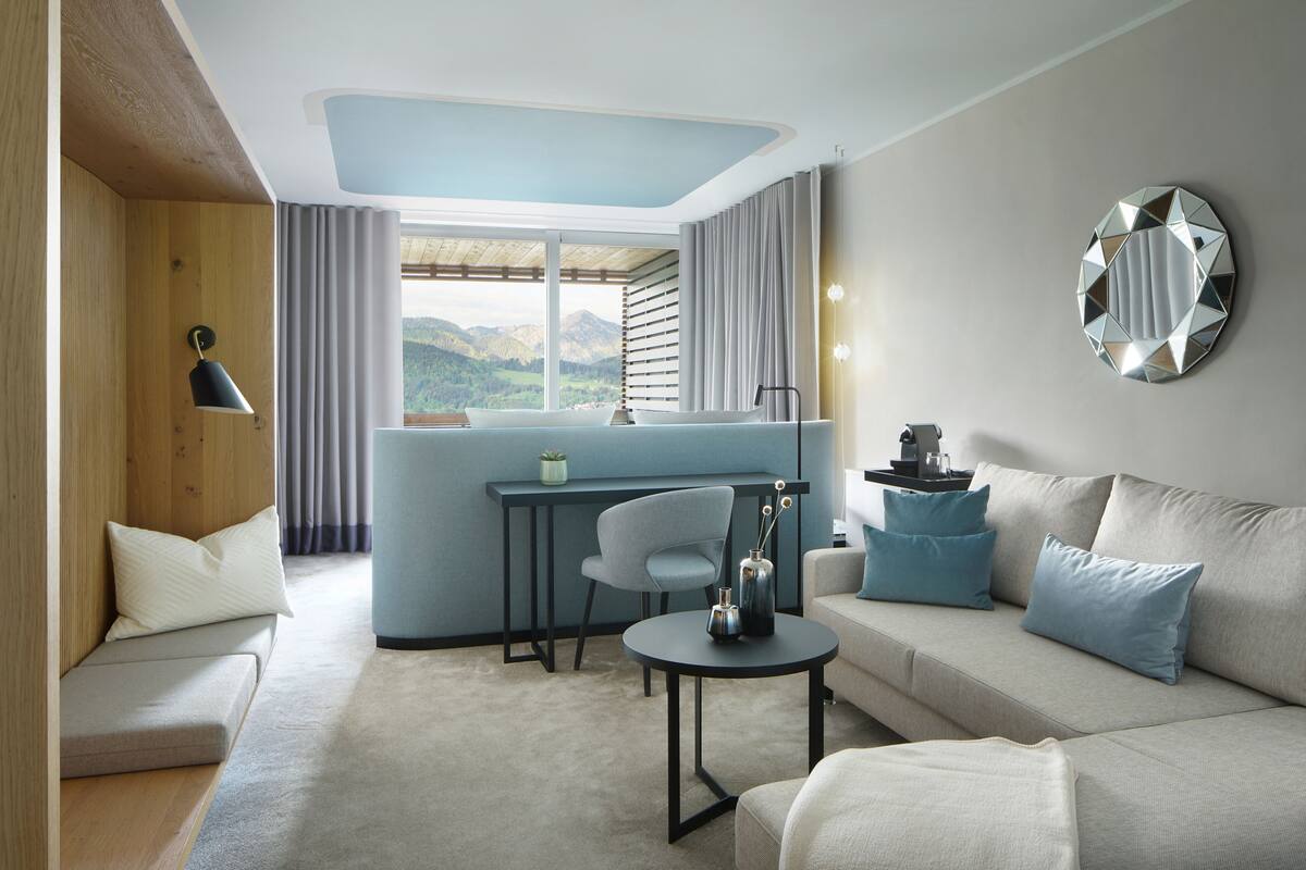 A modern hotel room with a sofa, a small table, a desk, and a view of the mountains.
