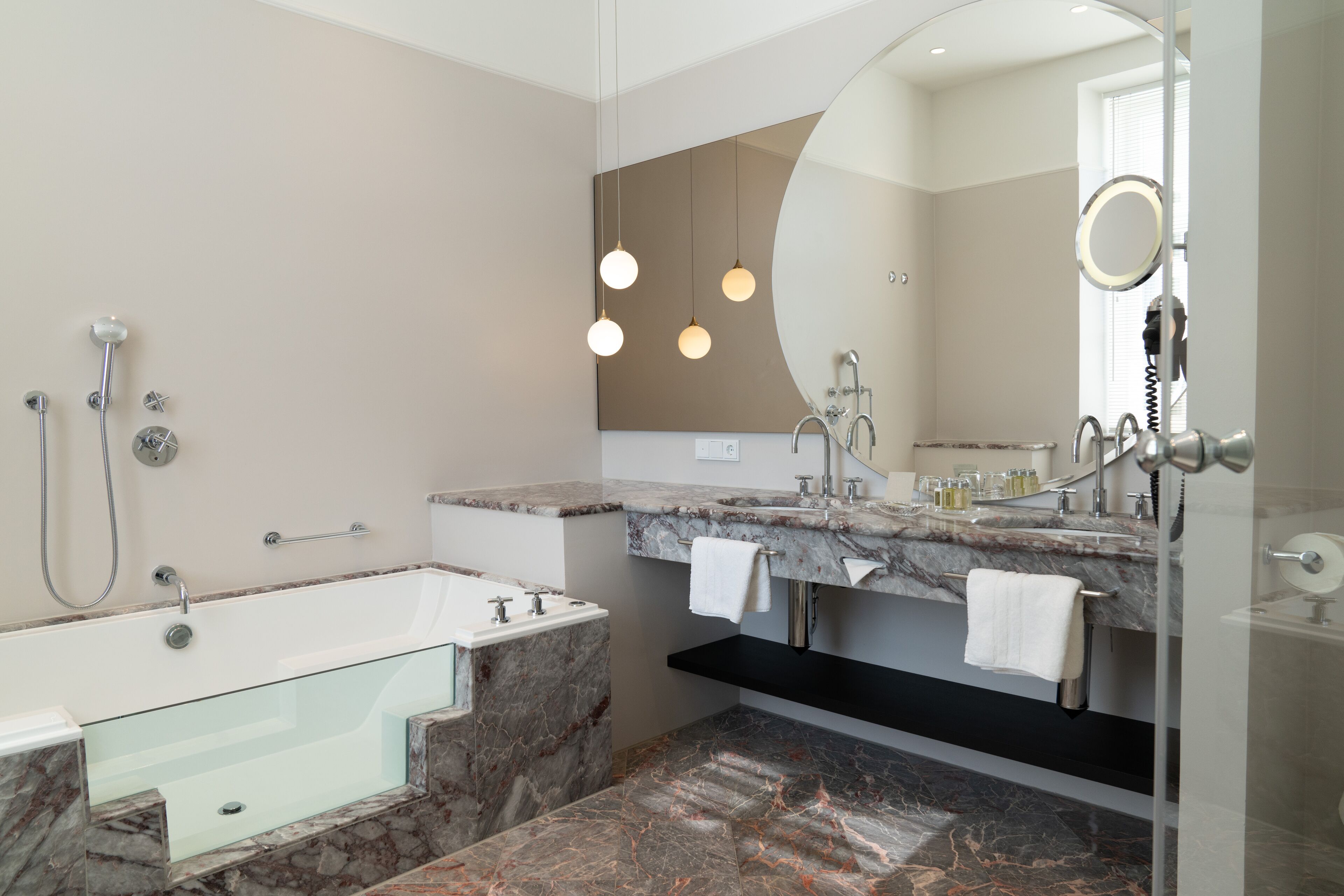 A modern bathroom with a glass-enclosed bathtub, a vanity with a marble countertop, and a large mirror with a round frame.