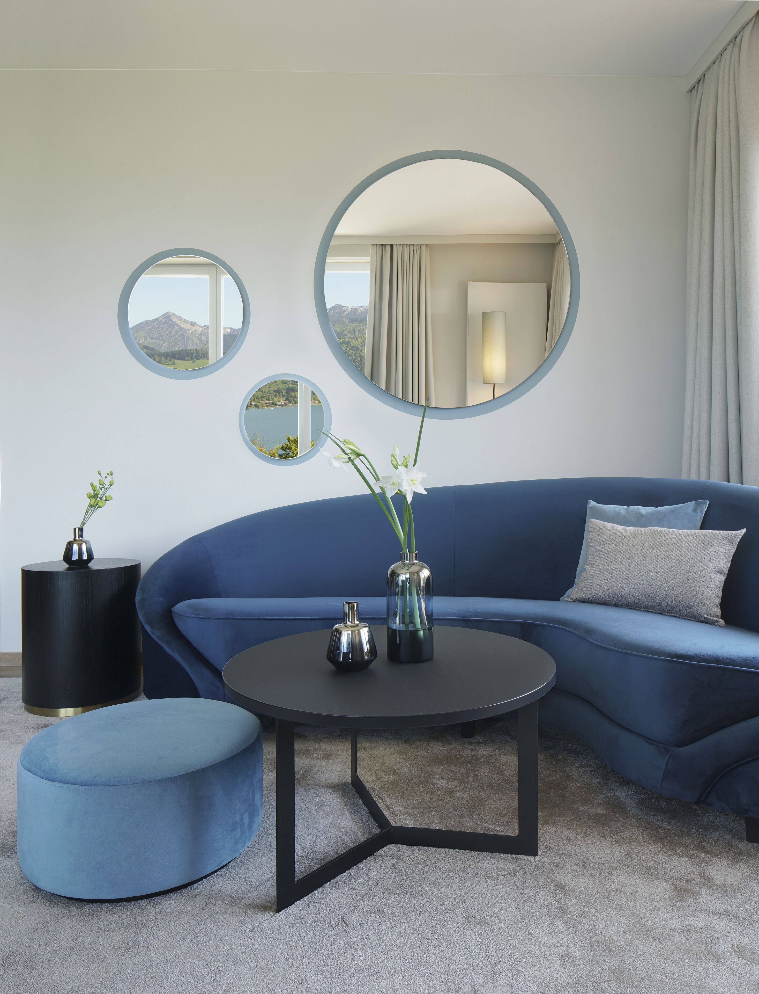 Ein modernes Wohnzimmer mit einem blauen Sofa, einem runden Couchtisch und zwei Vasen mit Blumen. Drei runde Spiegel hängen an der Wand, und durch einen davon ist eine Bergblick zu sehen.
