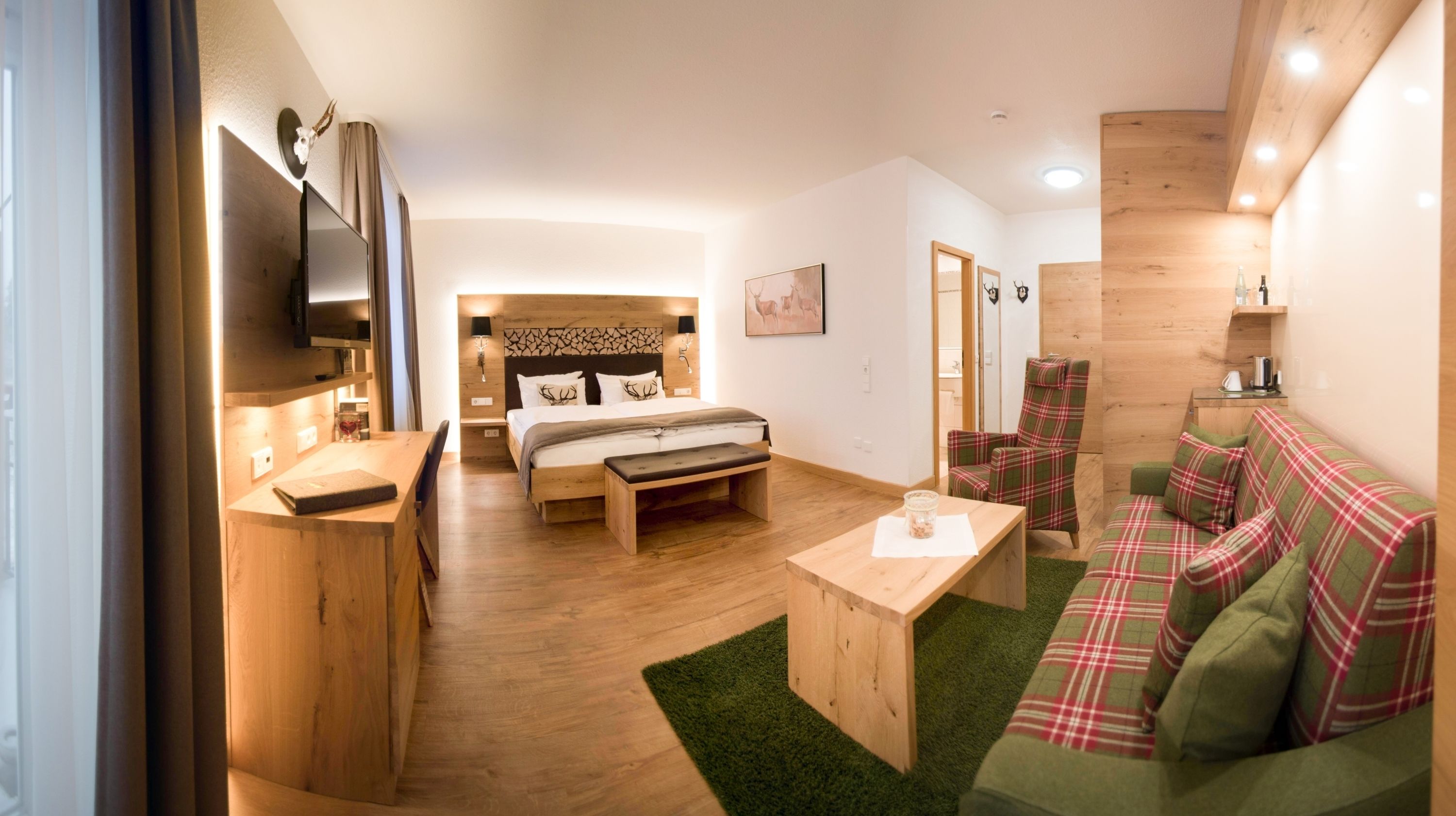 A modern hotel room with a wooden floor, a bed with a headboard, a green sofa, a wooden coffee table, and a TV mounted on the wall.