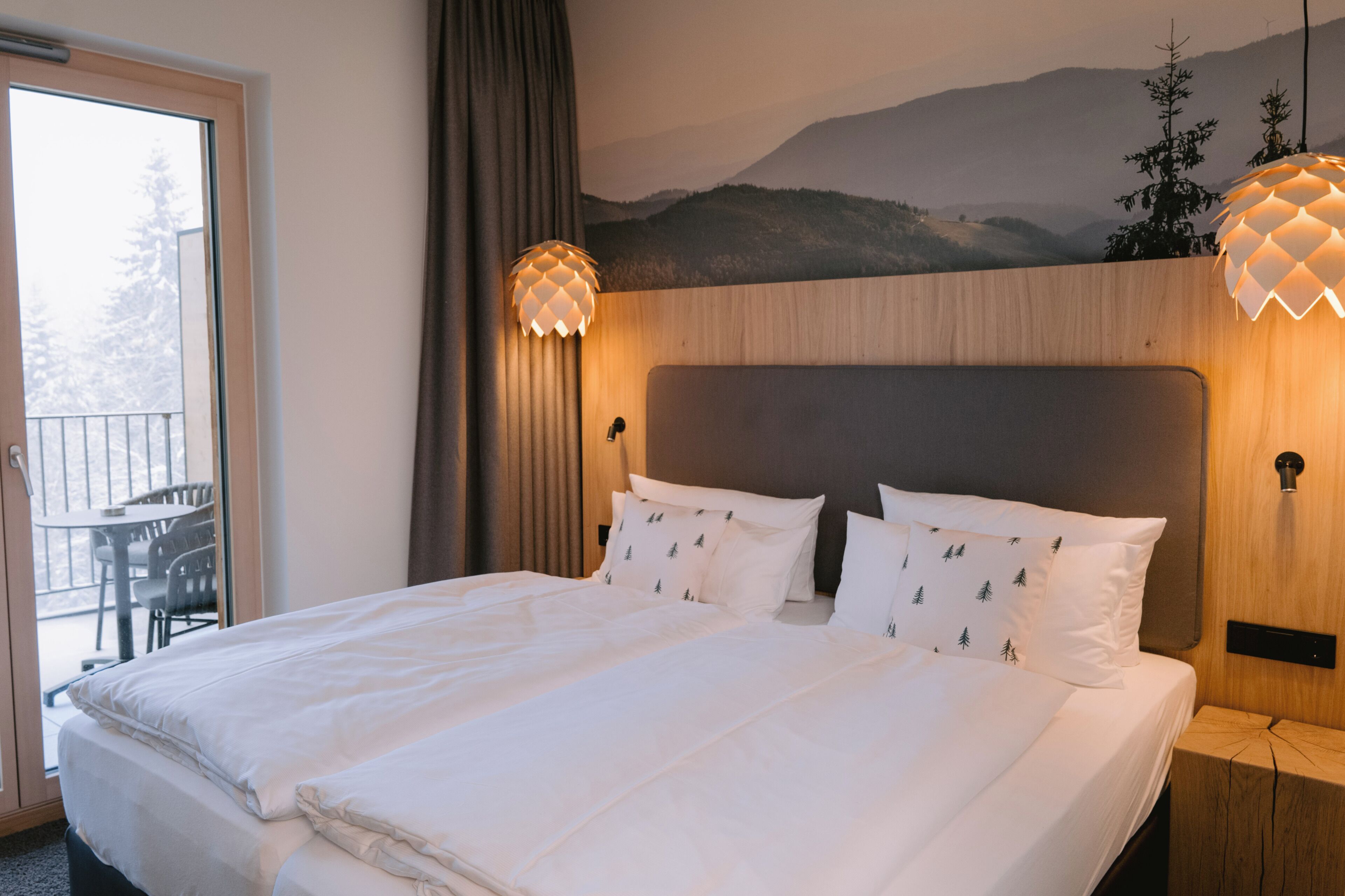 A hotel room with a bed, a view of mountains, a balcony with chairs, and a wooden headboard.