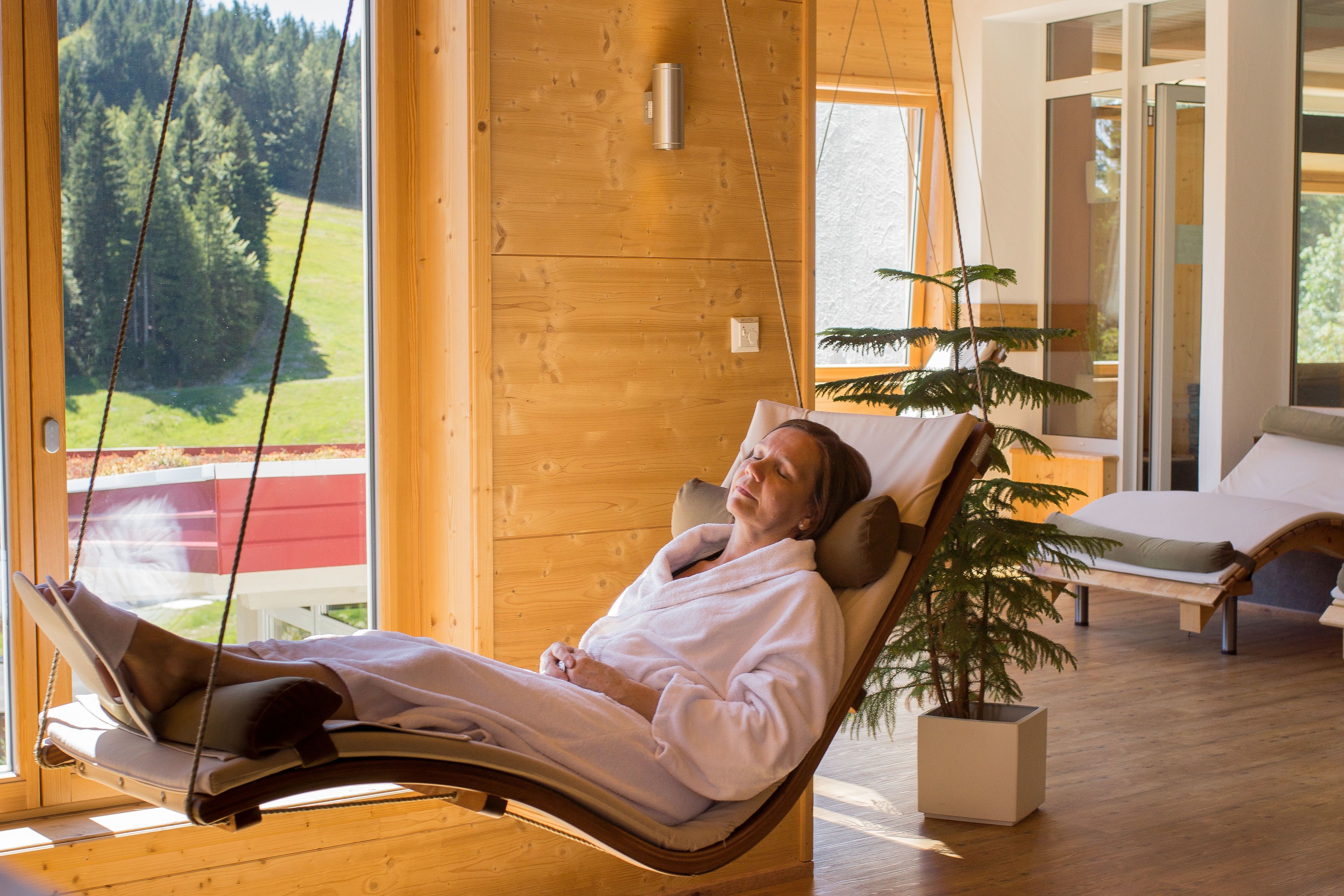 A person relaxing in a wooden cabin with a hammock chair, a bed, and a potted plant.