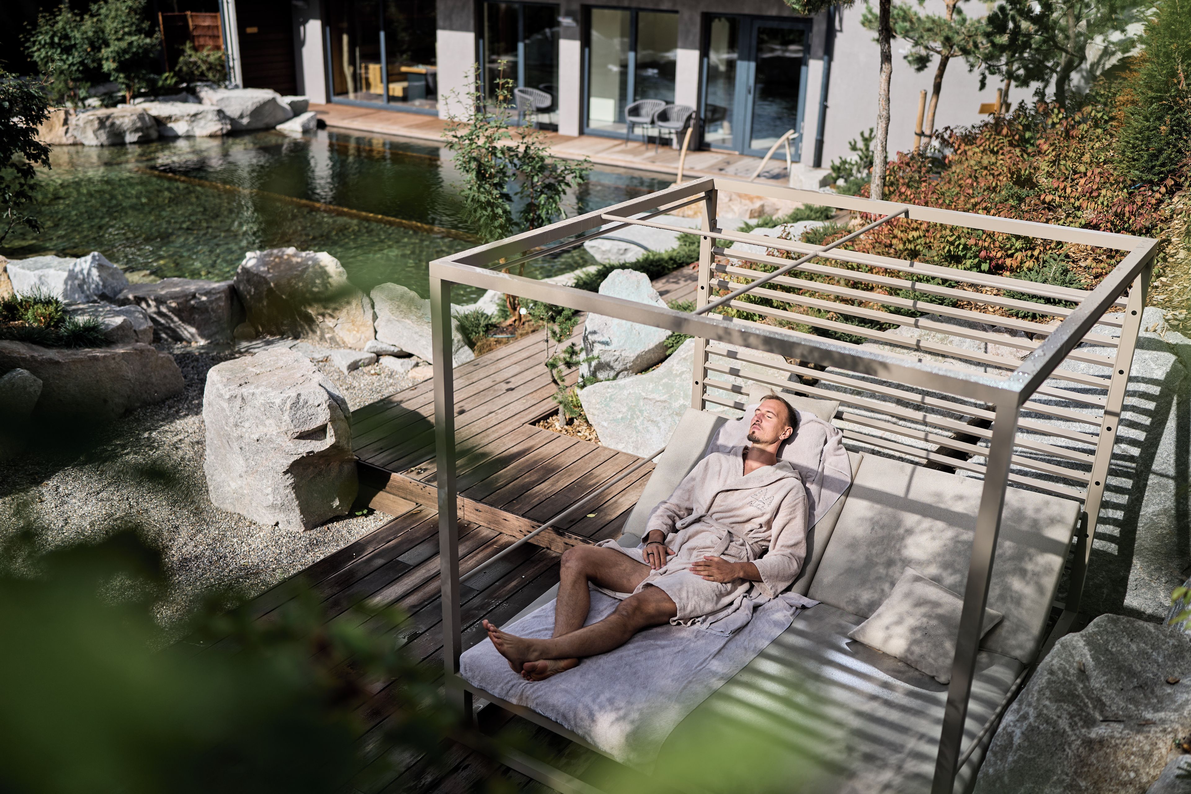 A person relaxing on a wooden deck by a pond with a pergola overhead.