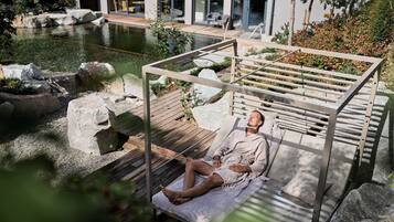 A person relaxing on a wooden deck by a pond with a pergola overhead.