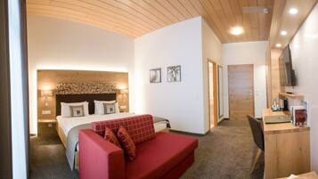 A modern hotel room with a wooden ceiling, a bed with a headboard, a red sofa, a desk with a chair, and a television.