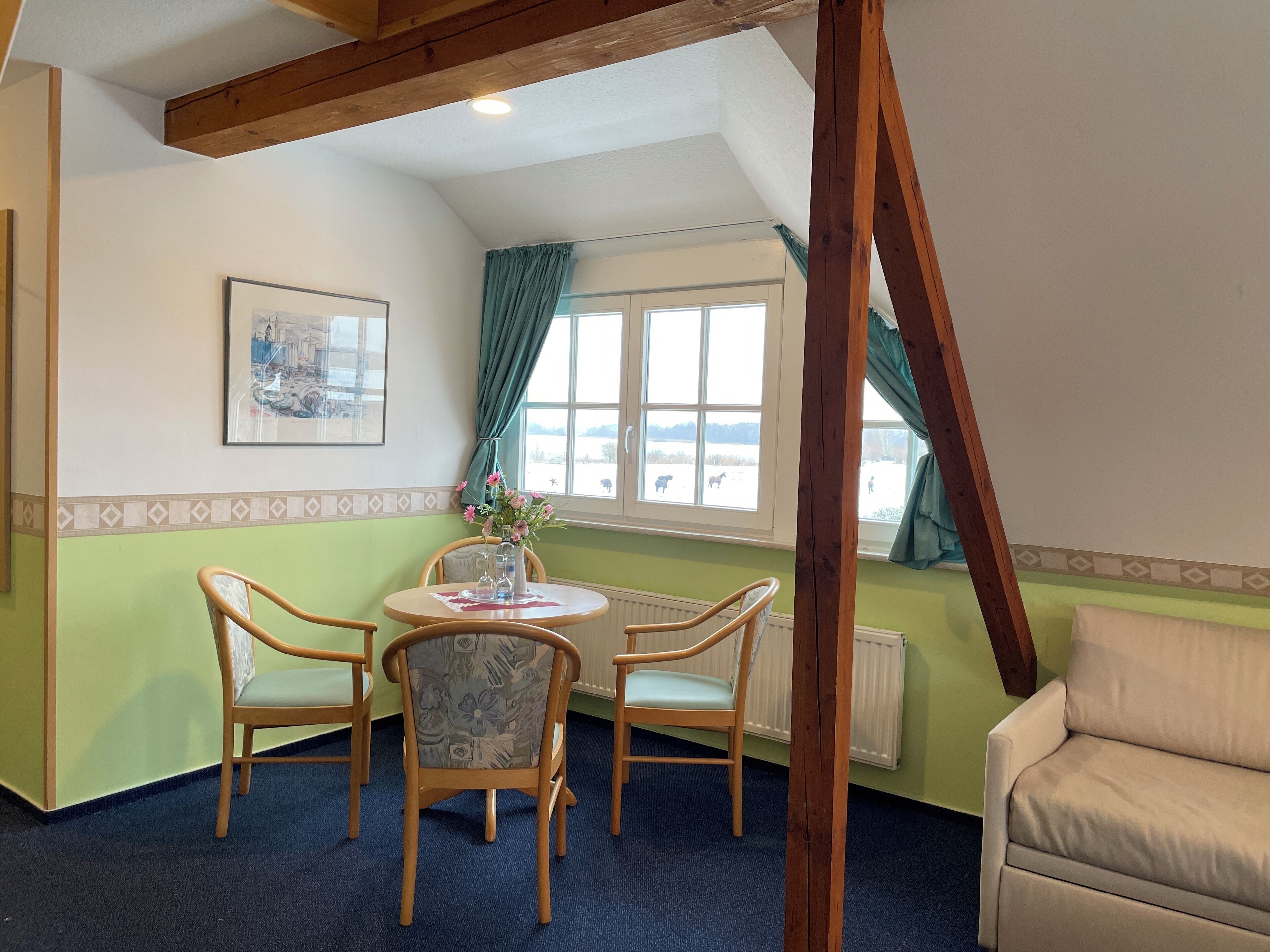 Ein Zimmer mit einem Tisch, vier Stühlen, einem Sofa, einem Fenster mit Vorhängen und einem Heizkörper.