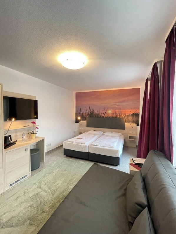 A hotel room with a bed, a desk with a computer, a TV, a painting of a sunset, and a window with curtains. - Flair Seehotel Zielow (Südmüritz)