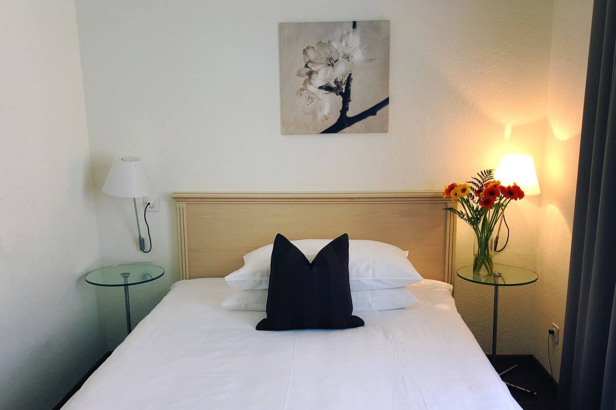 Cama bem arrumada com lençóis brancos e travesseiro preto, luminária fixada na parede, mesa de vidro com um vaso de flores e um quadro acima da cama.