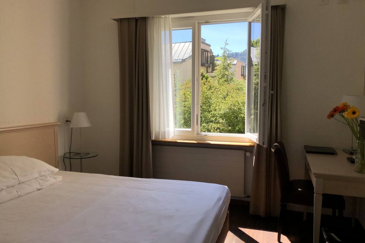 Quarto de hotel com cama, escrivaninha, cadeira, abajur e janela com cortinas.