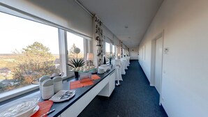 A hotel corridor with a long table set up for dining, large windows offering a view of trees and buildings, and a sign indicating Covid-19 safety measures. - Business - Hotel Artes (Chemnitz)
