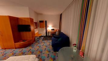 Ein Hotelzimmer mit einer Couch, einem an der Wand befestigten Fernseher, einem Schreibtisch mit einem Laptop und einem Bett mit weißer Bettdecke.