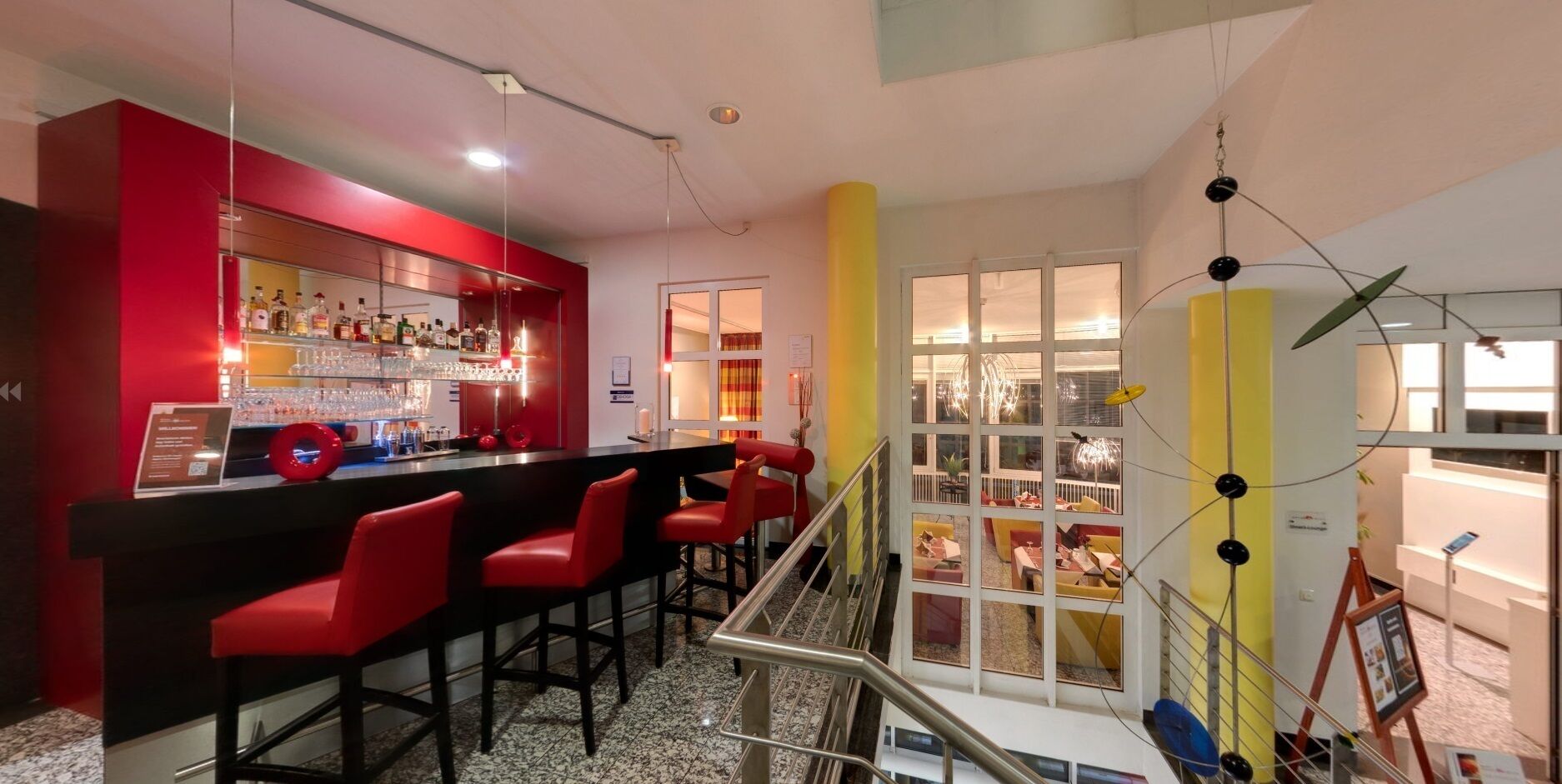 A bar area with red chairs and a bar counter, a staircase with a railing, and a view of the outdoor area through large windows.