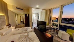 A hotel room with a bed, a TV, a sofa, a small table, and a window with a city view. - Business - Hotel Artes (Chemnitz)