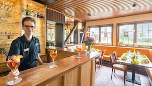Uma área de bar com balcão de madeira, copos e garrafas. Há também uma área de estar com mesas e cadeiras.