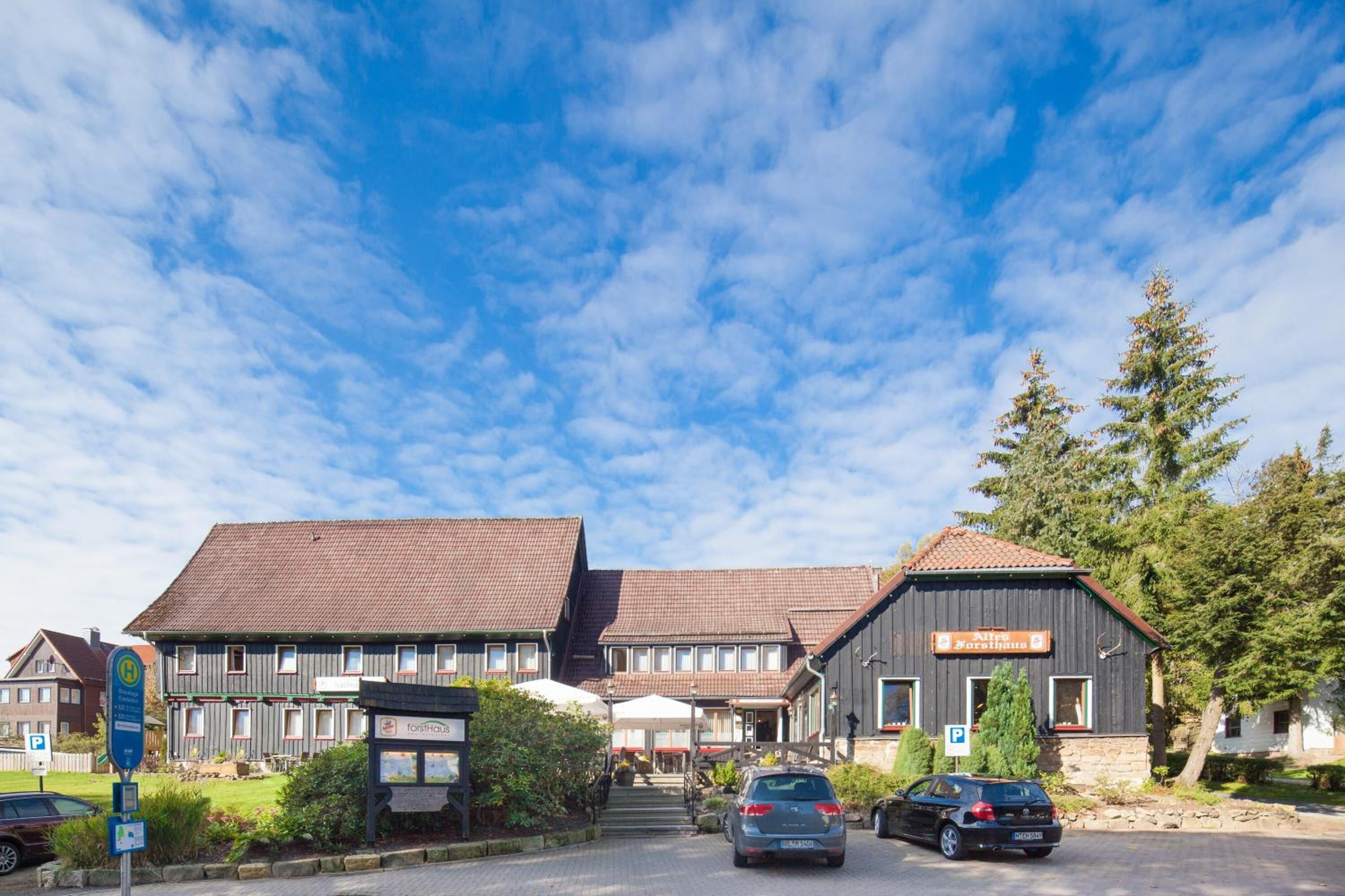 A hotel with a traditional architectural style, featuring a central building with a sloped roof and a smaller annex. There are cars parked in front, and a clear blue sky with scattered clouds above.