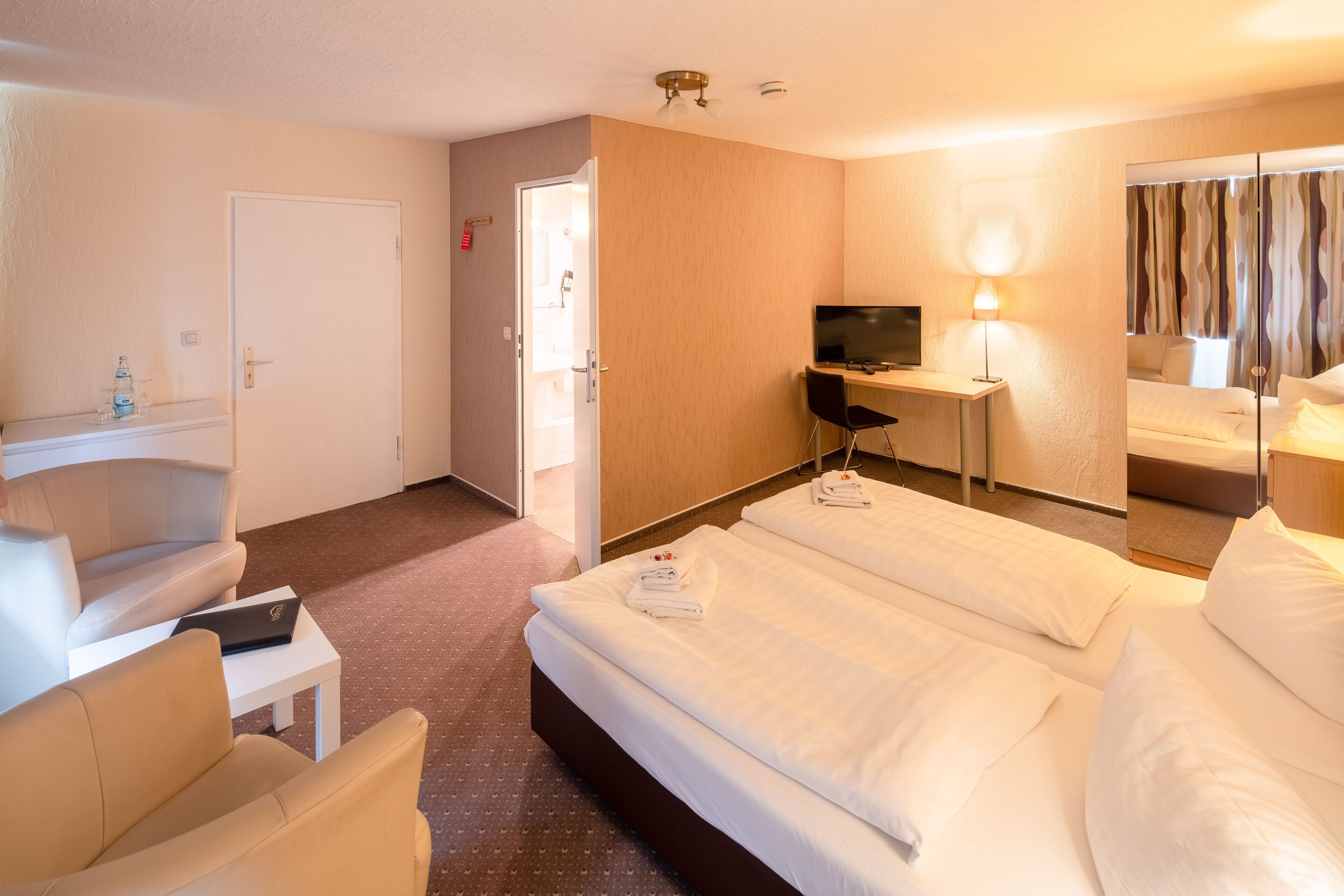 Camera d'albergo con due letti, una scrivania con un computer, una televisione e un bagno visibile attraverso una porta aperta.