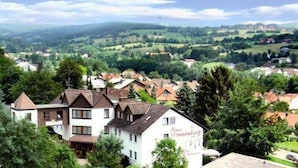 A white building with a sign reading "Ema Gmmbirg" surrounded by lush greenery and a hilly landscape. - AKZENT Hotel Haus Sonnenberg (Schotten)