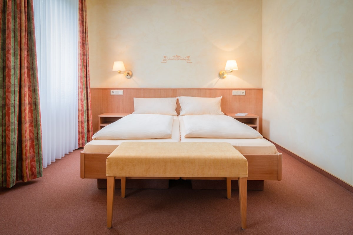 Uma cama de casal com cabeceira de madeira, duas mesas de cabeceira, um banquinho e um aplique de parede.