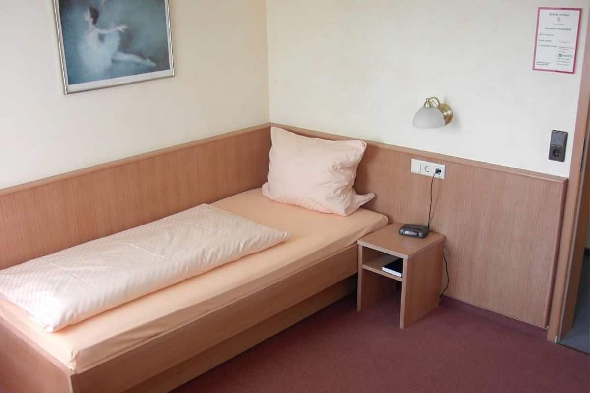 Uma cama de solteiro com cabeceira de madeira, uma mesa de cabeceira com abajur e uma tomada na parede.