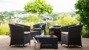 An outdoor seating area with wicker furniture, a table with a bottle of wine, and a view of a town in the background. - AKZENT Hotel Haus Sonnenberg (Schotten)