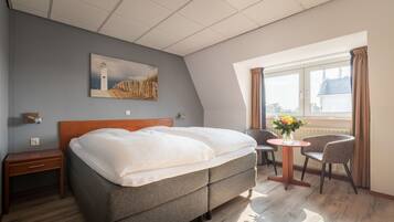 A bedroom with a bed, a nightstand, a chair, a small table with flowers, and a window with curtains.
