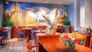 A restaurant with a mural of a lighthouse, wooden tables, chairs, and a vase of flowers on the table. - Hotel Astoria (Noordwijk)