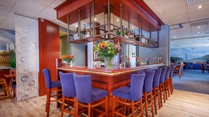 A bar area with a wooden bar counter, blue bar stools, and a decorative flower arrangement. - Hotel Astoria (Noordwijk)