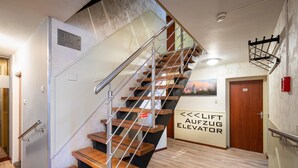 A modern staircase with wooden steps and metal railings, a sign indicating an elevator, and a wall-mounted light fixture. - Hotel Astoria (Noordwijk)