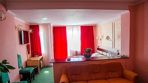Una habitación con sofá, escritorio y silla, una mesita con un florero y ventana con cortinas rojas.