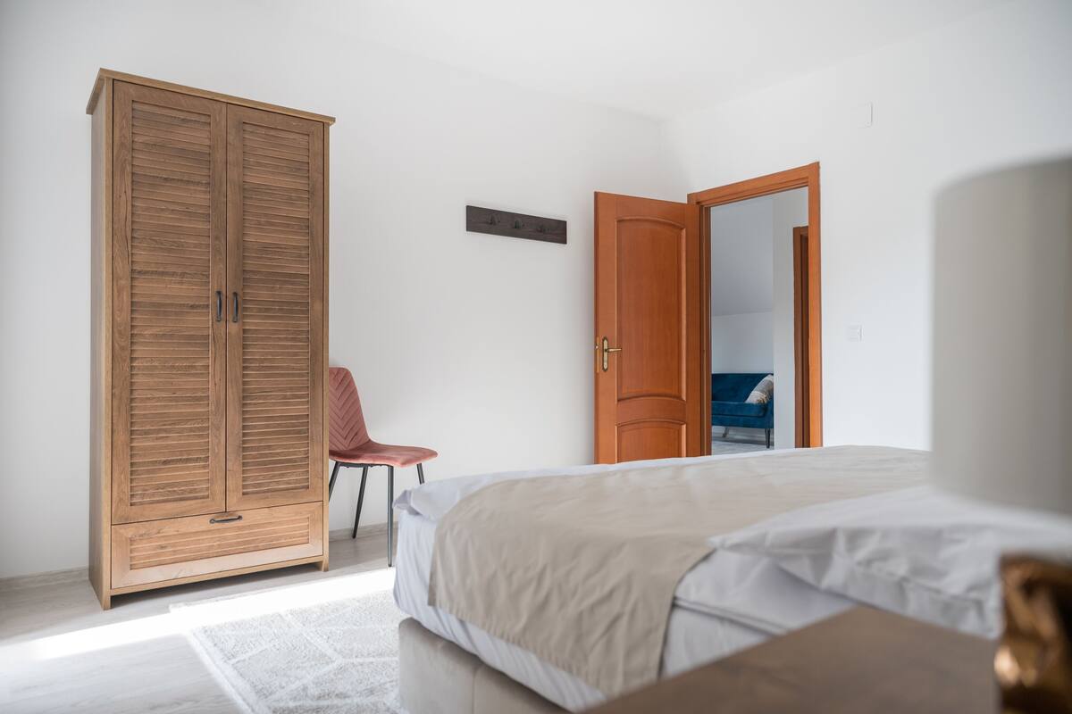 A bedroom with a bed, a wooden wardrobe, a red chair, and a door leading to another room.