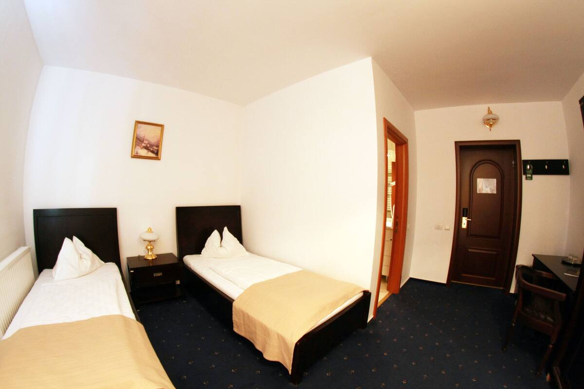 A hotel room with two single beds, a small table, a chair, a door, and a wall-mounted light fixture.