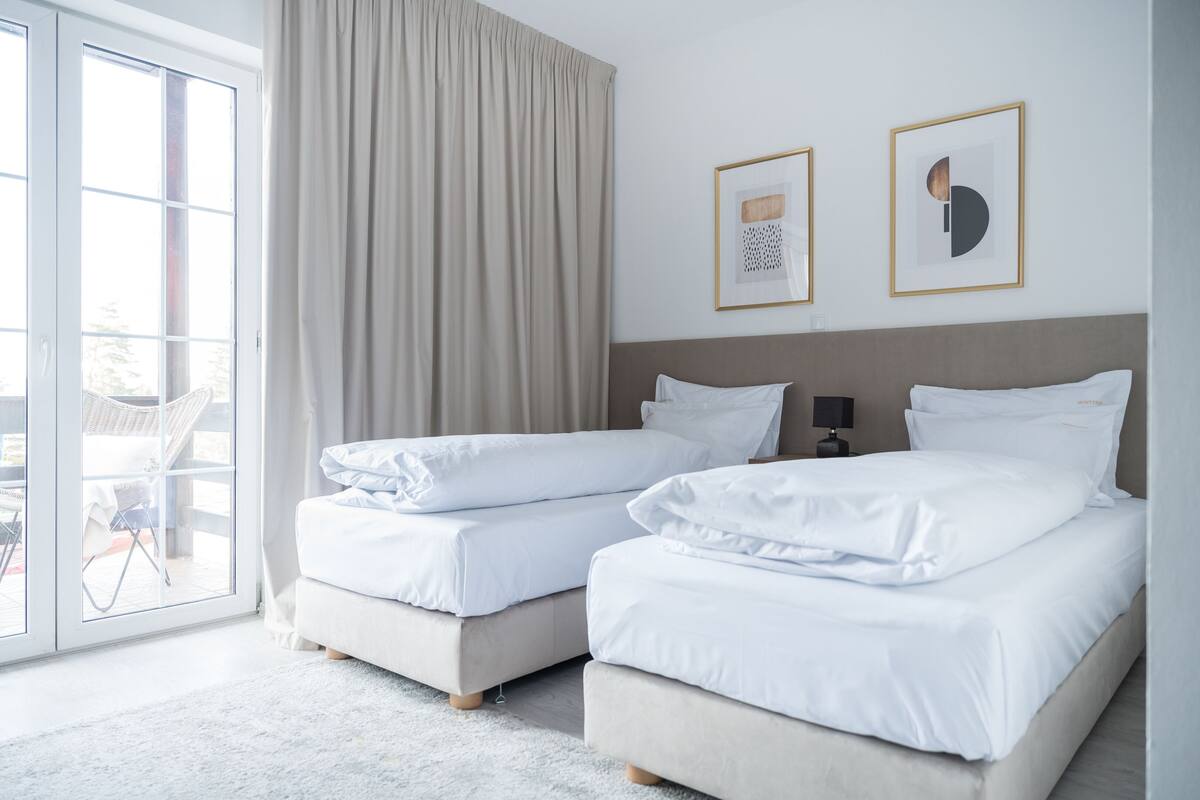 A hotel room with two beds, a large window with curtains, and two framed pictures on the wall.