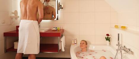 A man standing at a bathroom sink with a towel wrapped around his waist, a woman relaxing in a bathtub filled with rose petals, and a mirror reflecting the scene.