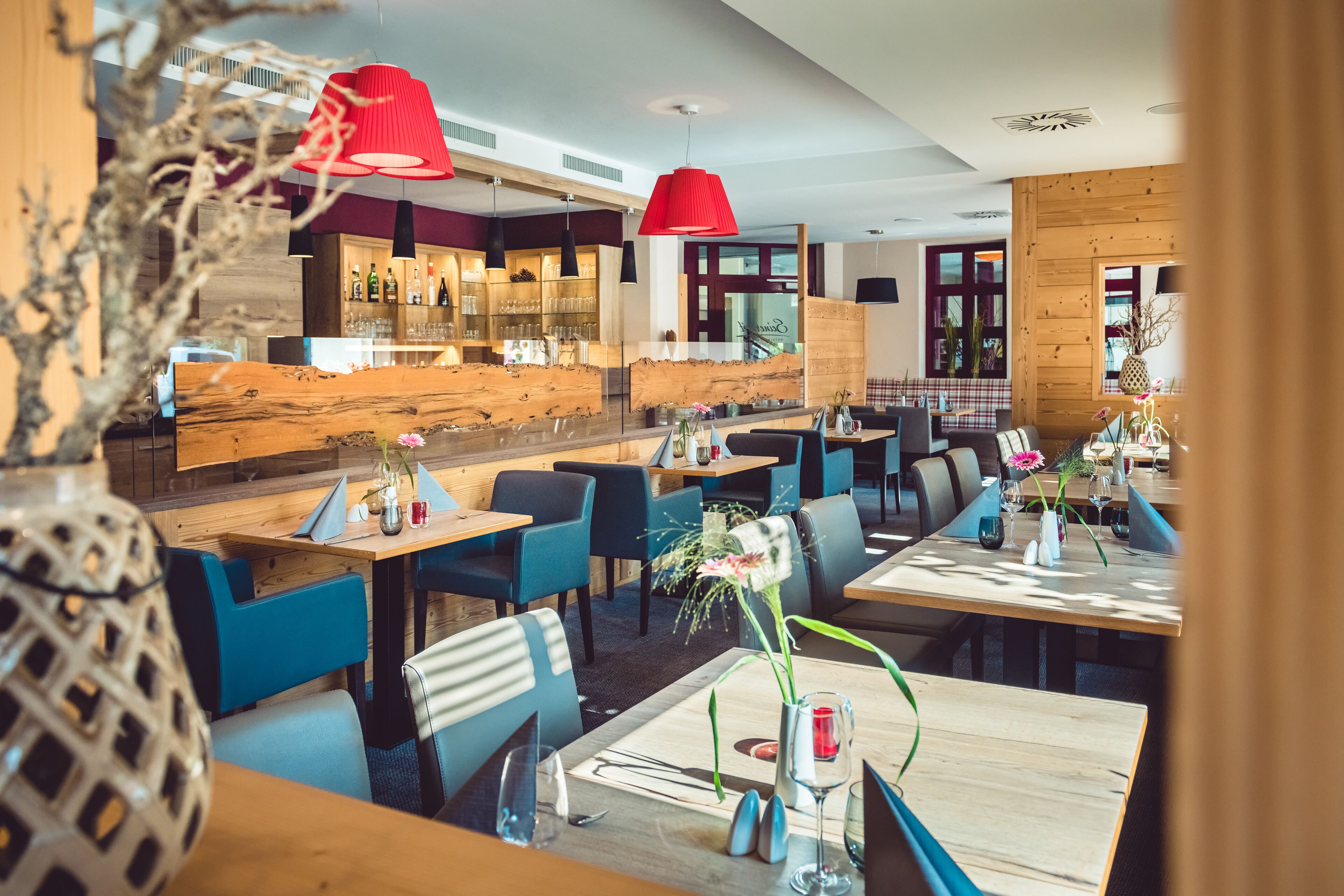 Un restaurante moderno con mesas de madera y sillas azules, lámparas colgantes rojas y una zona de bar al fondo.