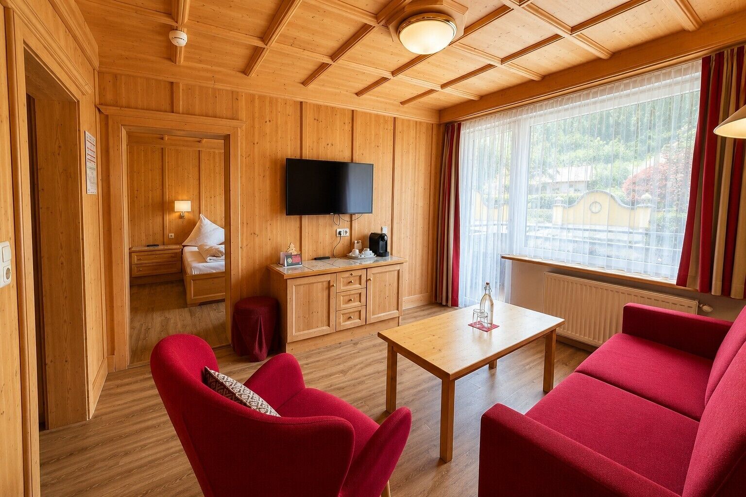 A wooden-paneled room with a bed, a TV, a table, and red chairs.