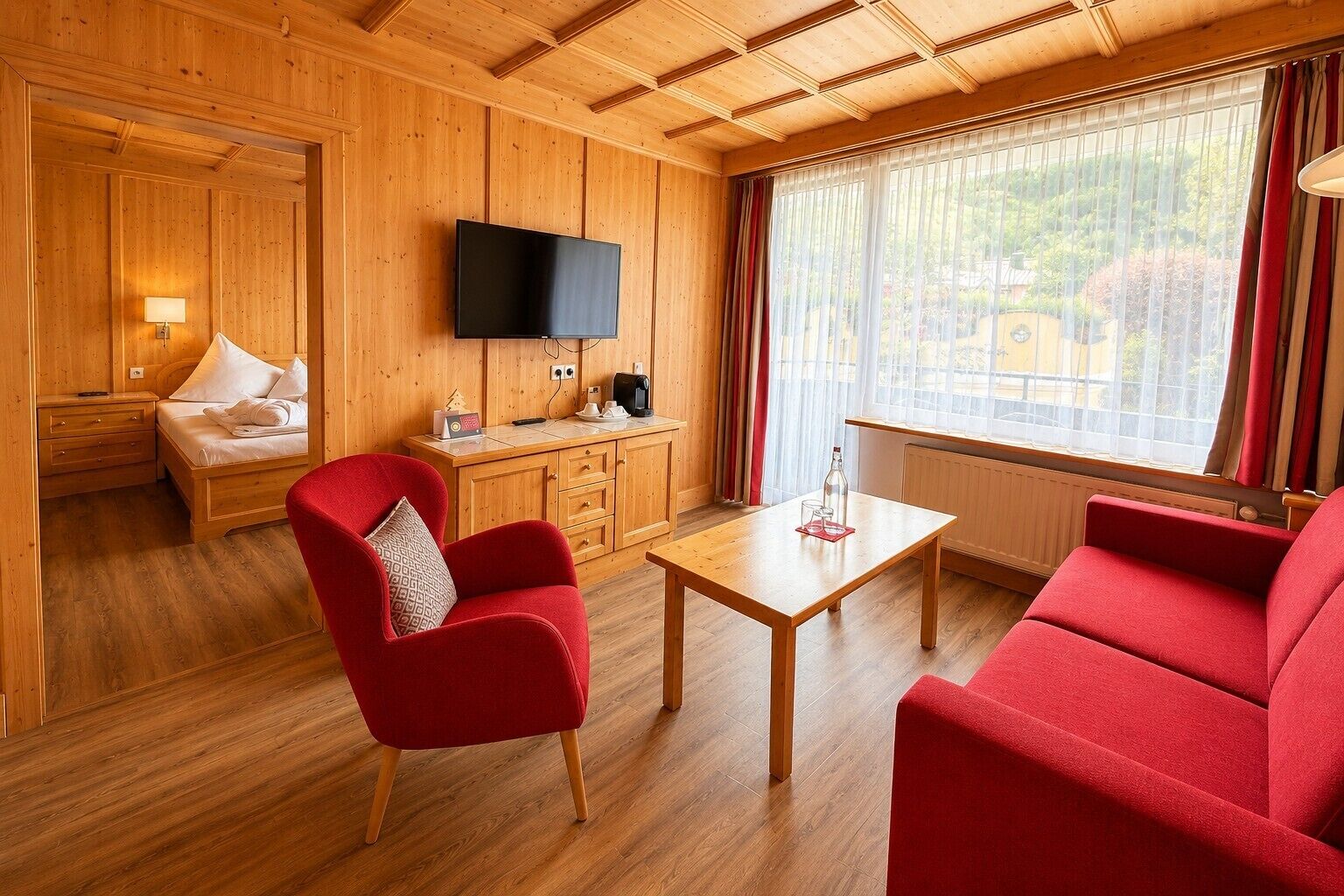 Una habitación con cama, televisión, una silla roja, una mesa de madera y una ventana con cortinas.