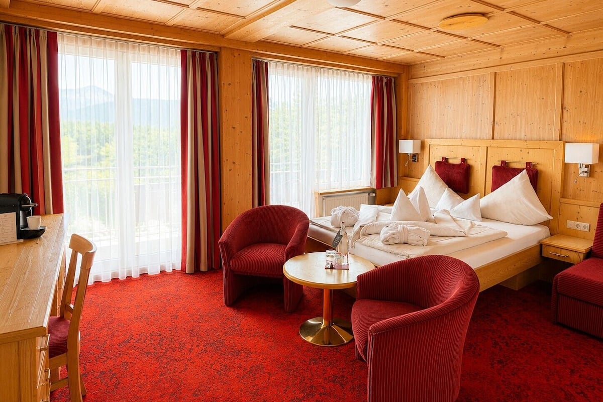 A hotel room with a wooden ceiling, a bed with white linens, a red upholstered chair, a small round table, and a view of the outdoors through large windows.