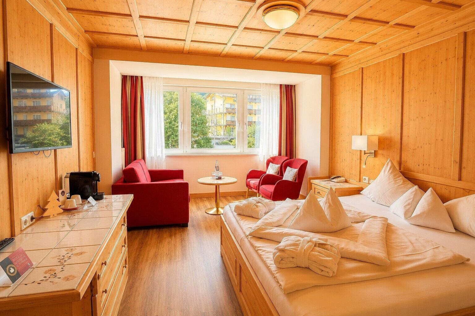 Een hotelkamer met houten inrichting, een bed met witte lakens, een rode bank, een klein rond tafeltje en een televisie aan de muur.