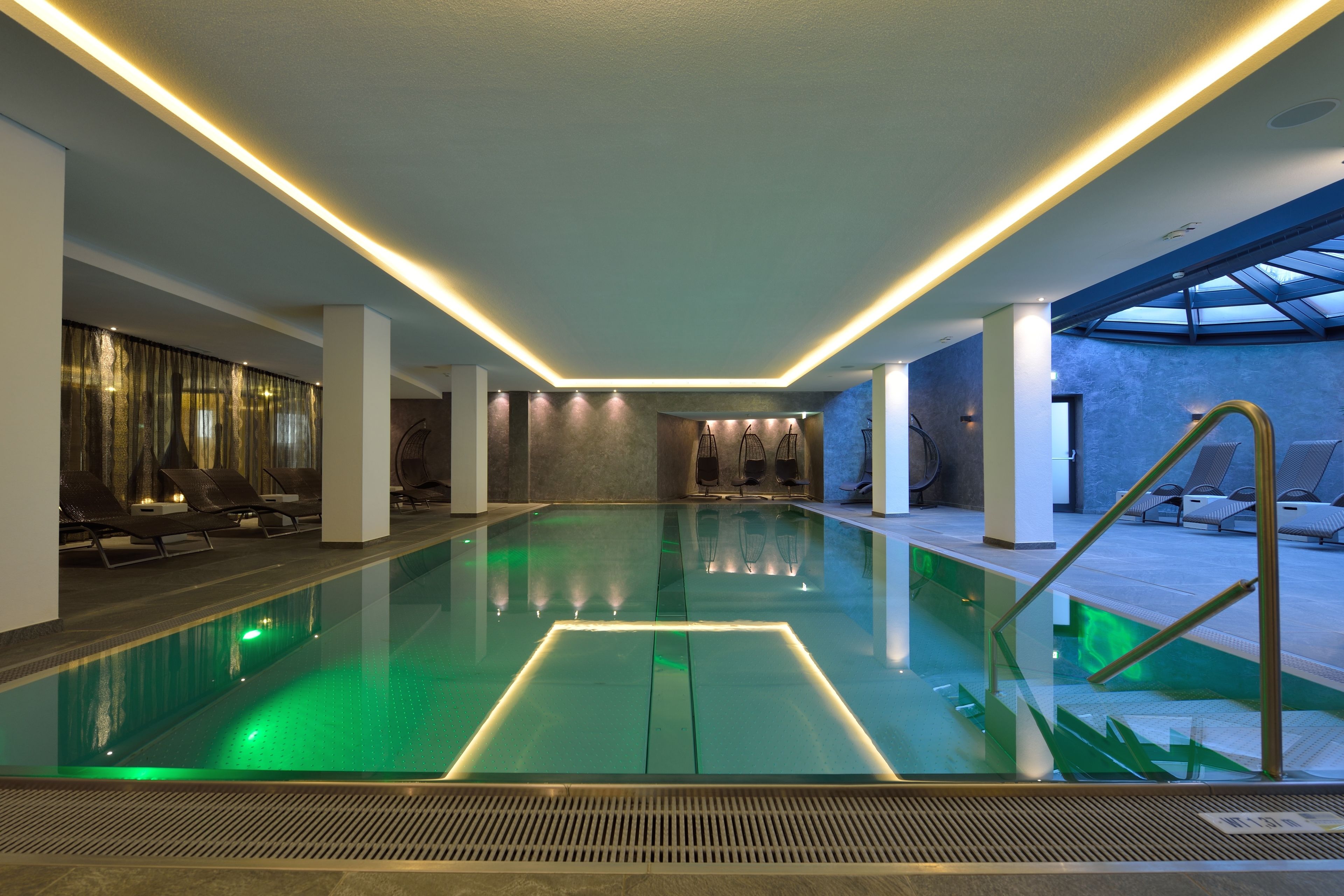 Une piscine intérieure moderne avec un fond en verre, entourée de chaises longues et dotée d’une verrière.