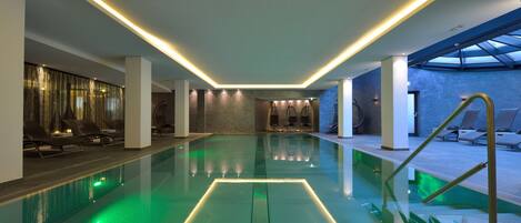 A modern indoor swimming pool with a glass bottom, surrounded by lounge chairs and a skylight.