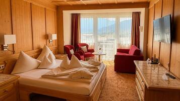 Quarto de hotel com cama, duas poltronas vermelhas, uma mesinha, televisão e varanda com cortinas.