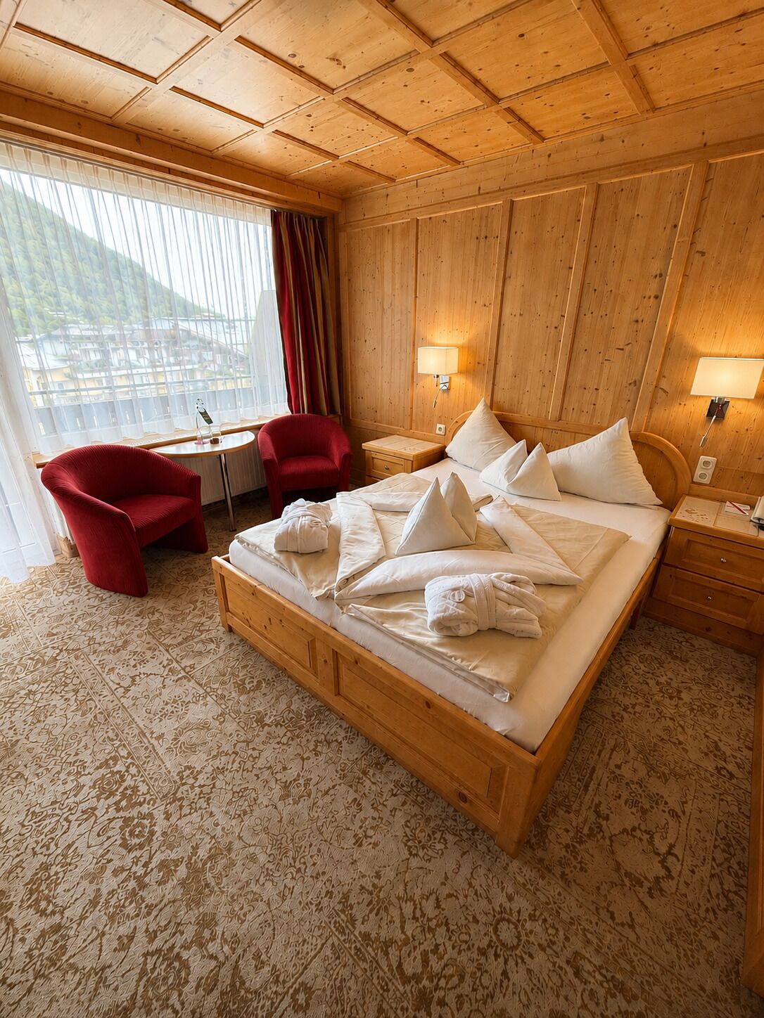 A hotel room with a wooden bed, a red armchair, a small table, and a view of a mountain.