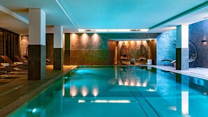 Una piscina interior moderna con agua cristalina, sillas de descanso y un área de spa que incluye una silla de masaje y una pequeña mesa.