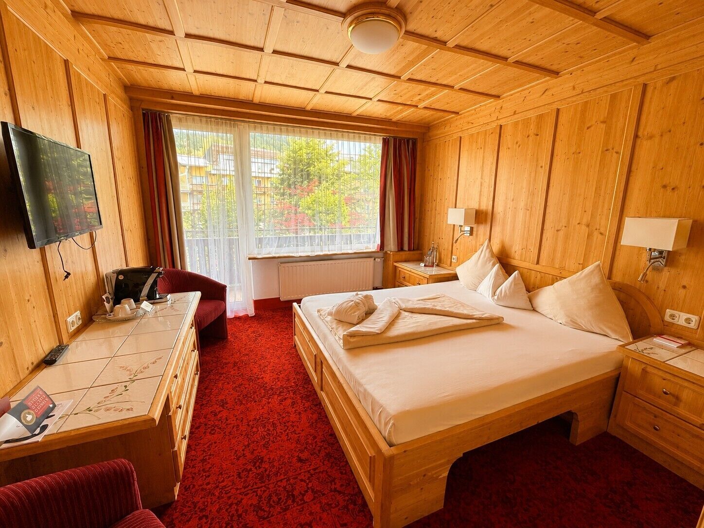 A hotel room with a wooden interior, a bed with pillows, a desk with a chair, a television, and a window with curtains.