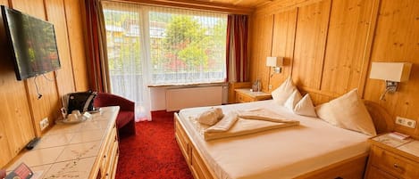 Ett hotellrum med träinredning, en säng med kuddar, ett skrivbord med en stol, en tv och ett fönster med gardiner.