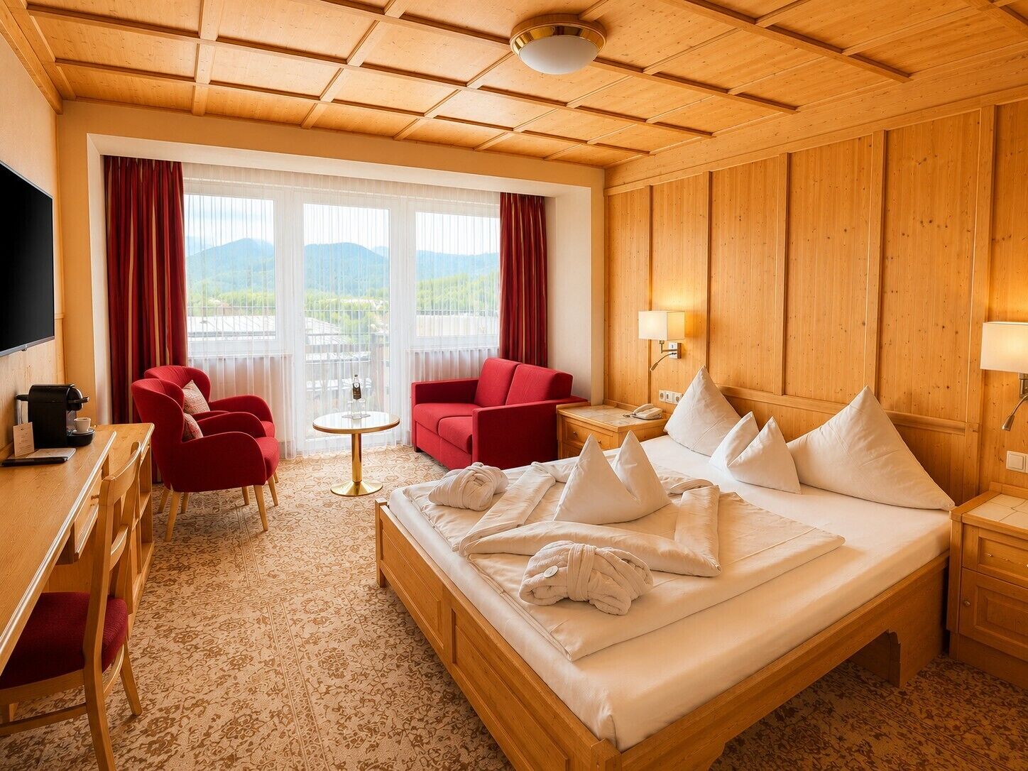 A hotel room with a bed, two red armchairs, a small round table, a desk with a coffee maker, and a TV.