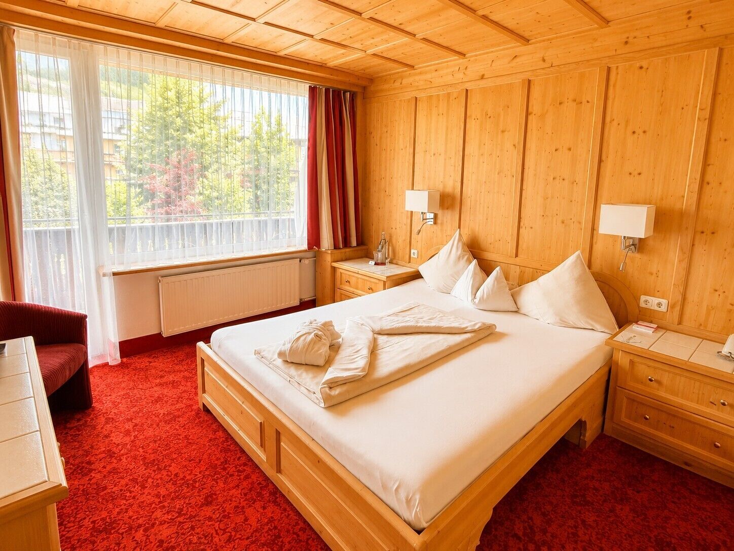 A hotel room with a wooden bed, two bedside tables, a red carpet, a window with curtains, and a view of trees.