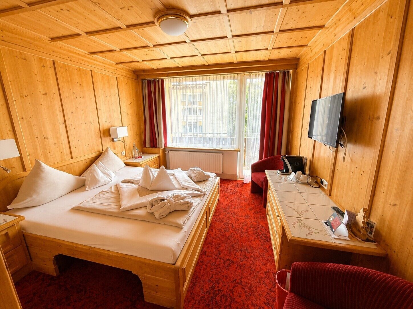 Habitación de hotel con techo de madera, una cama con sábanas blancas, un escritorio con ordenador, una silla y un televisor.