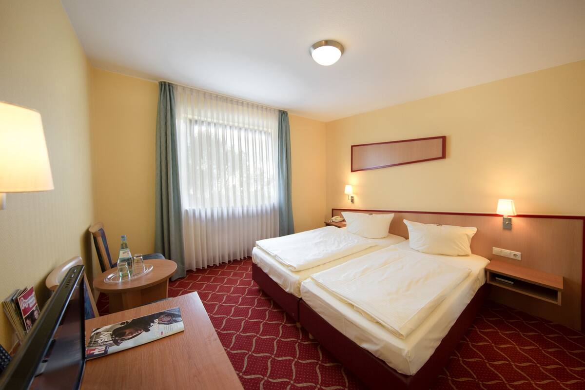 A hotel room with two beds, a desk with a chair, a small table with a bottle and glasses, and a window with curtains.